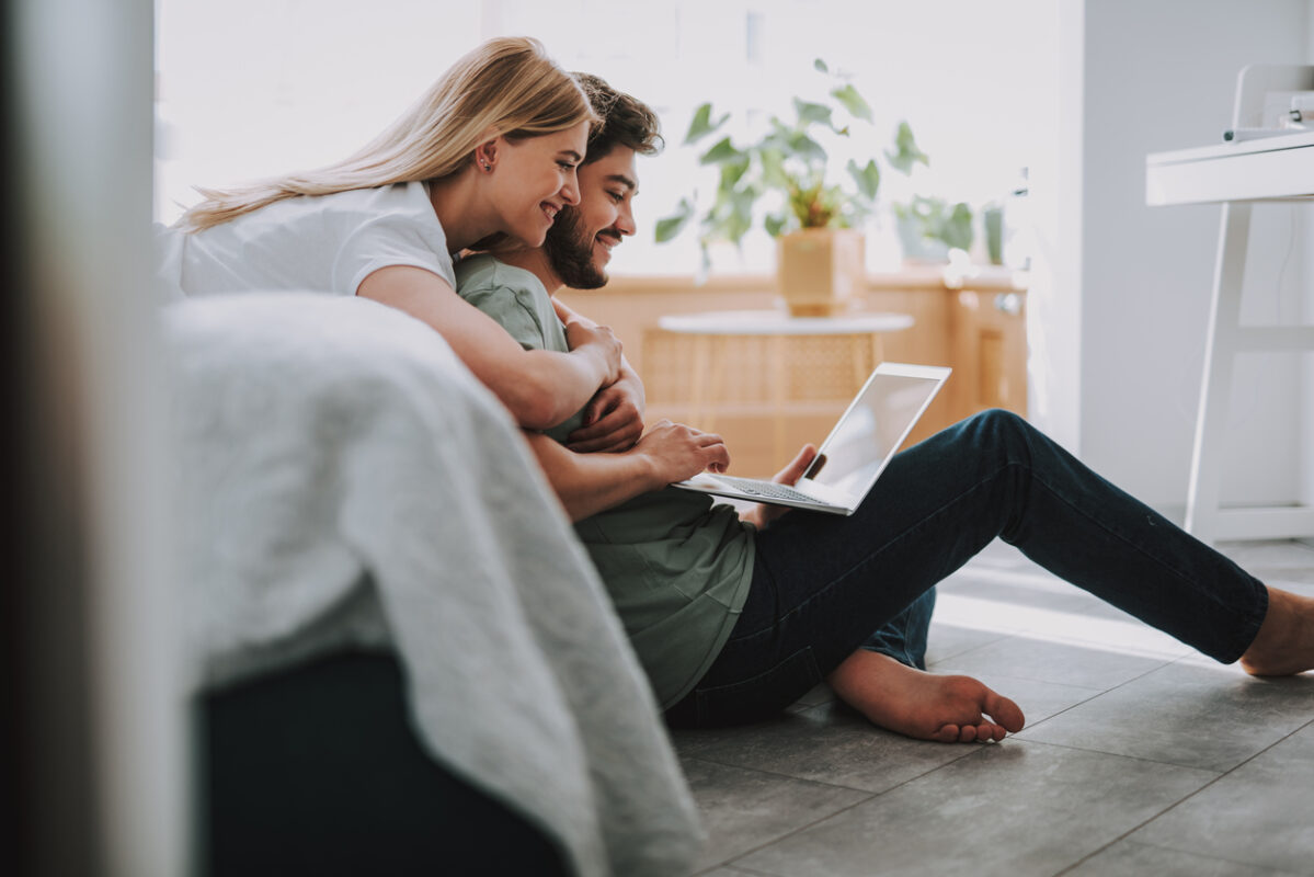 Cheerful beautiful woman embracing her bearded husband while resting together in the bedroom looking at laptop for mortgage