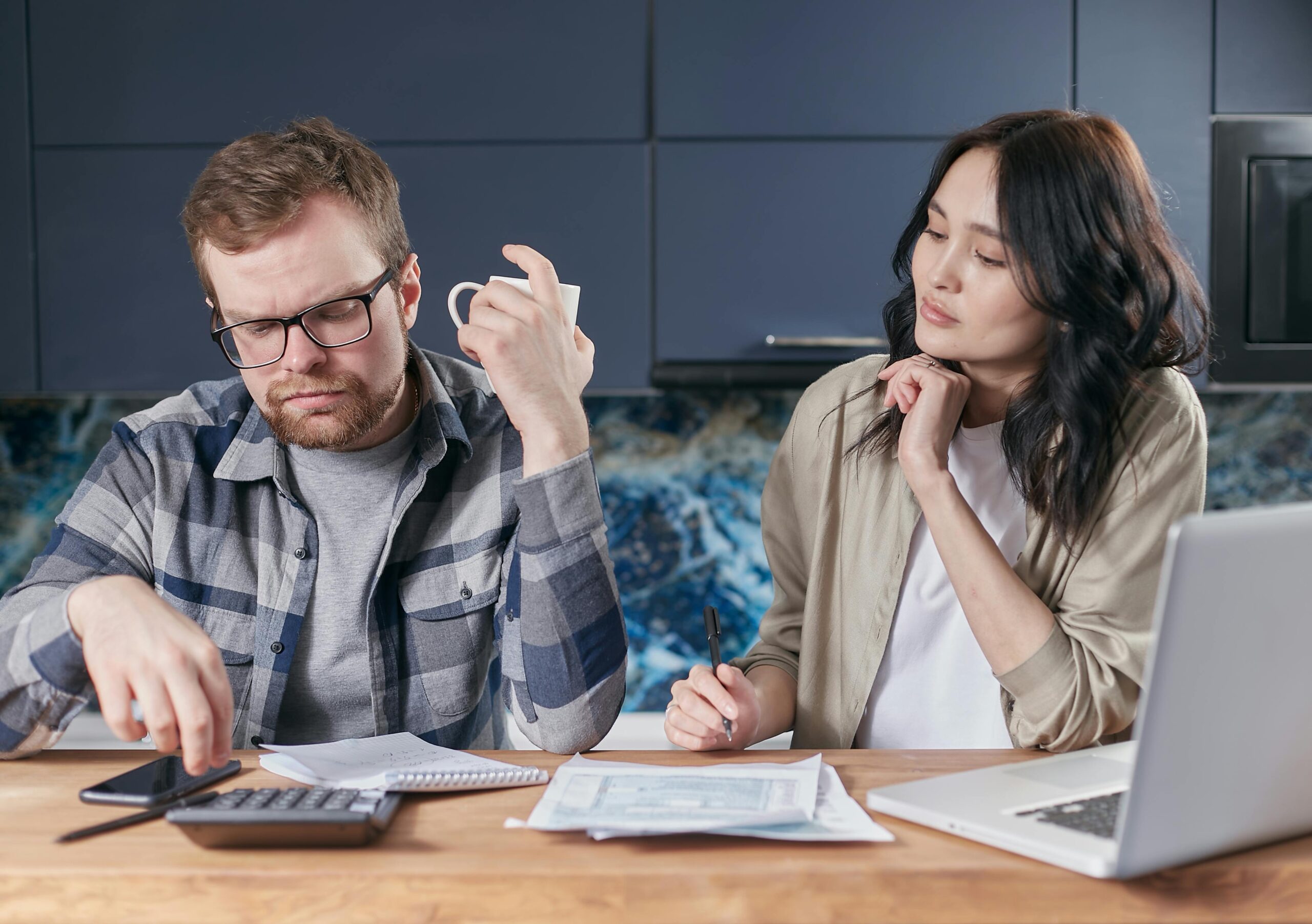 young to middle aged male and female couple sat at desk with paperwork, calculator and laptop looking serious