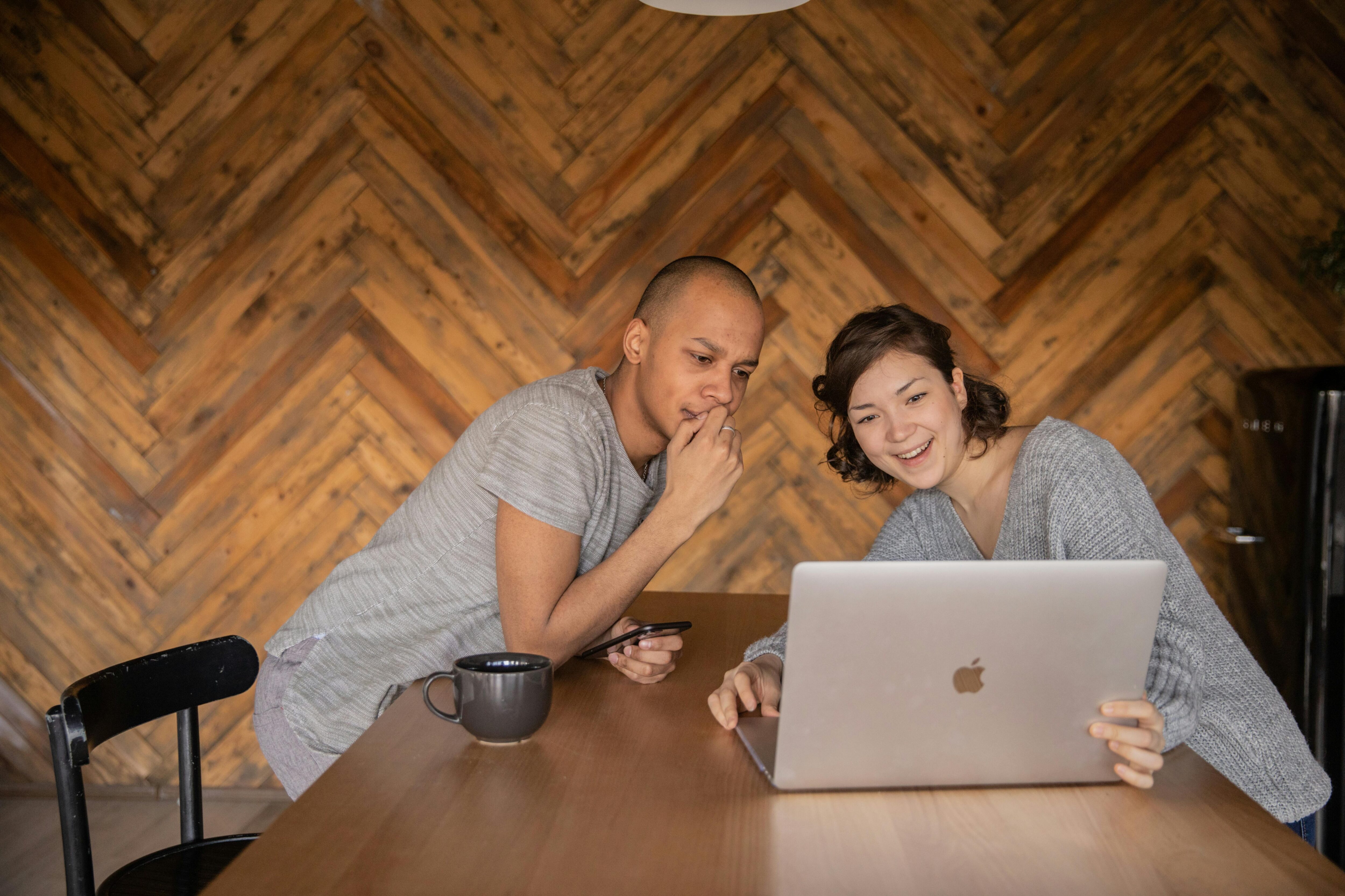 Happy mixed race man and woman couple sat at table looking at a laptop