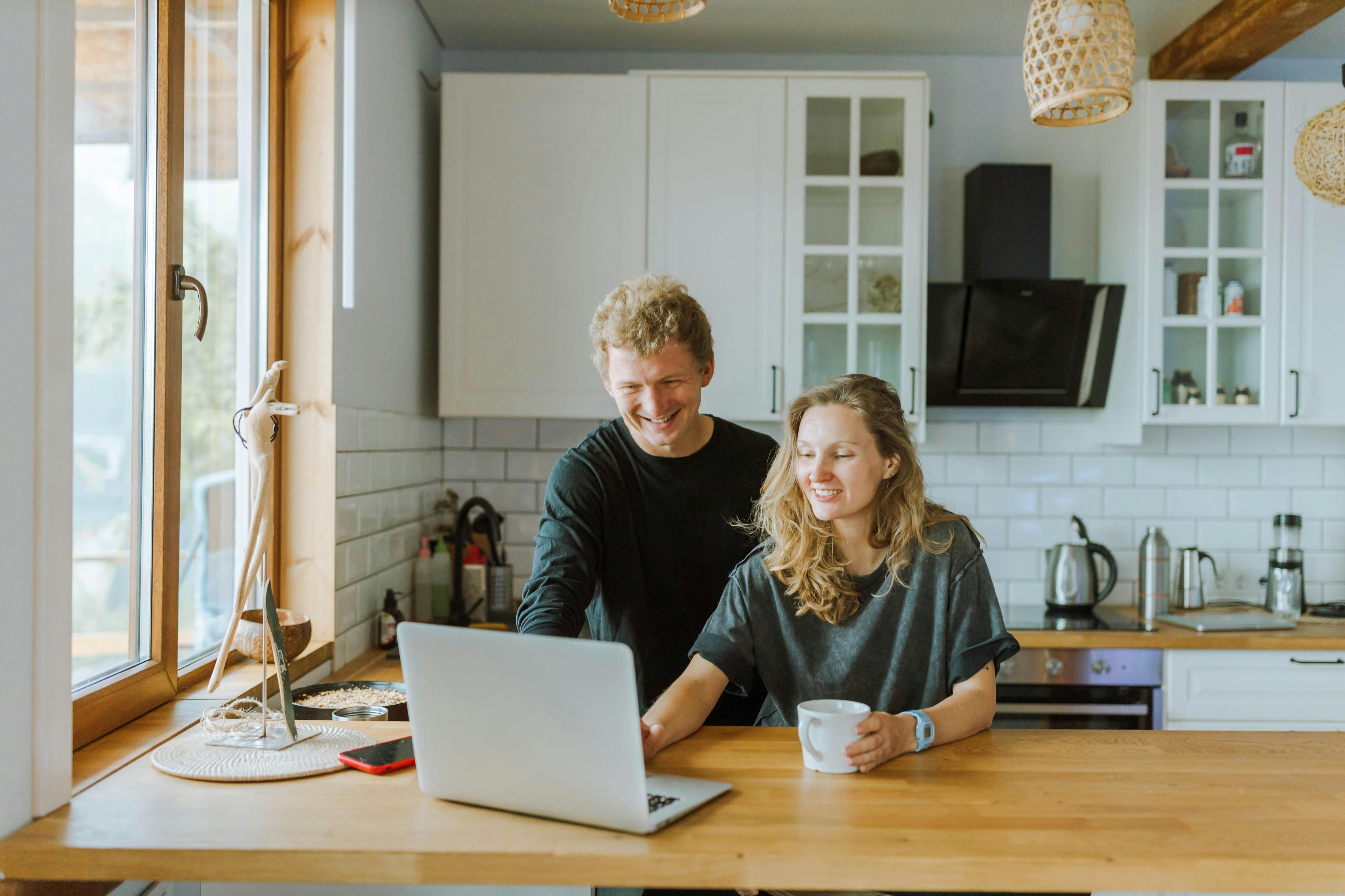 Happy male and female couple in the kitchen with a hot drink and a laptop on the kitchen work top